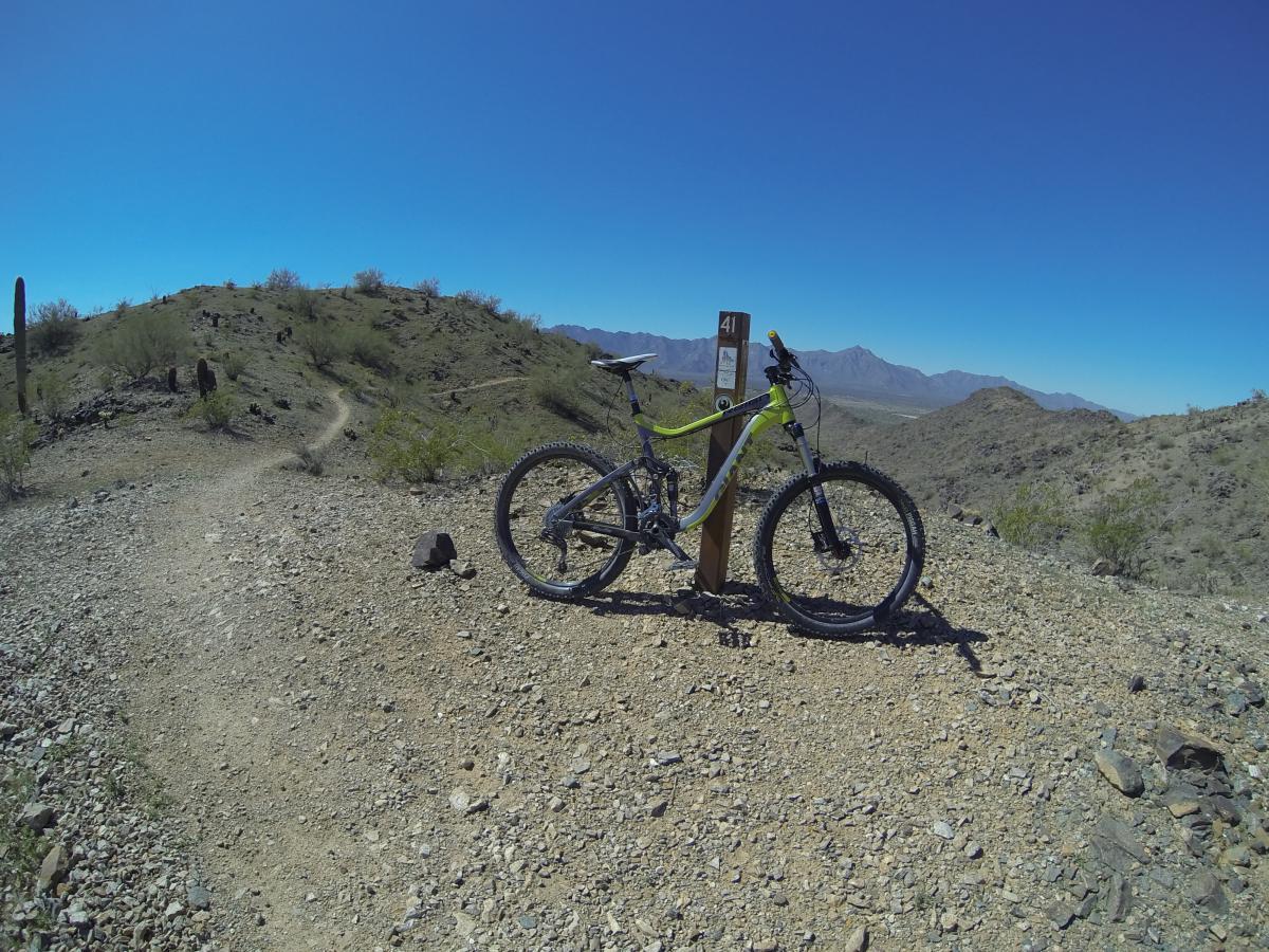 A mountain bike resting against a trail marker on a rocky path, with a clear blue sky and distant mountains in the background. South Mountain Park / National Trail mountain bike trail.