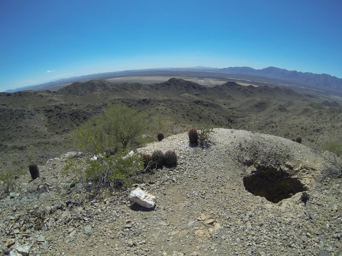A wide view of a rocky desert landscape under a clear blue sky, featuring rolling hills and scattered vegetation. The foreground includes a rocky pathway, with cacti and shrubs. In the distance, mountains are visible against the horizon, and a flat valley stretches out below. South Mountain Park / National Trail mountain bike trail.