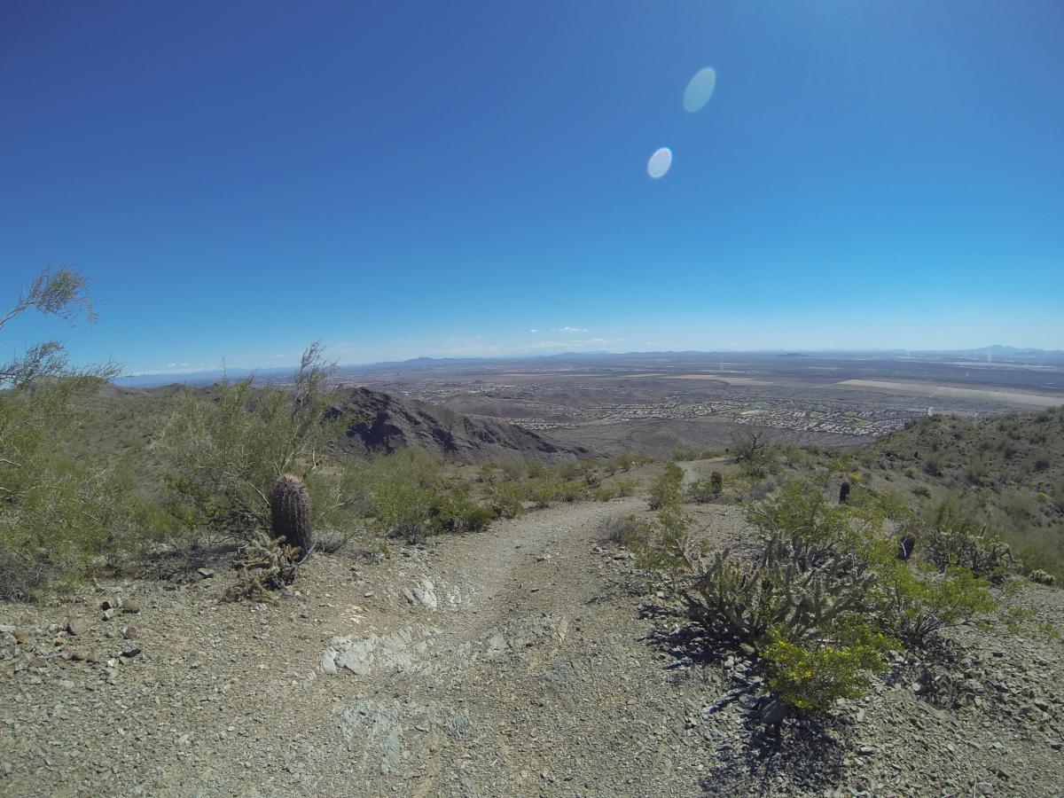 A panoramic view of a desert landscape from a mountain trail, featuring rocky terrain, scattered vegetation, and a clear blue sky. In the distance, the horizon includes rolling hills and signs of human settlement, with patches of greenery visible in the valley below. South Mountain Park / National Trail mountain bike trail.