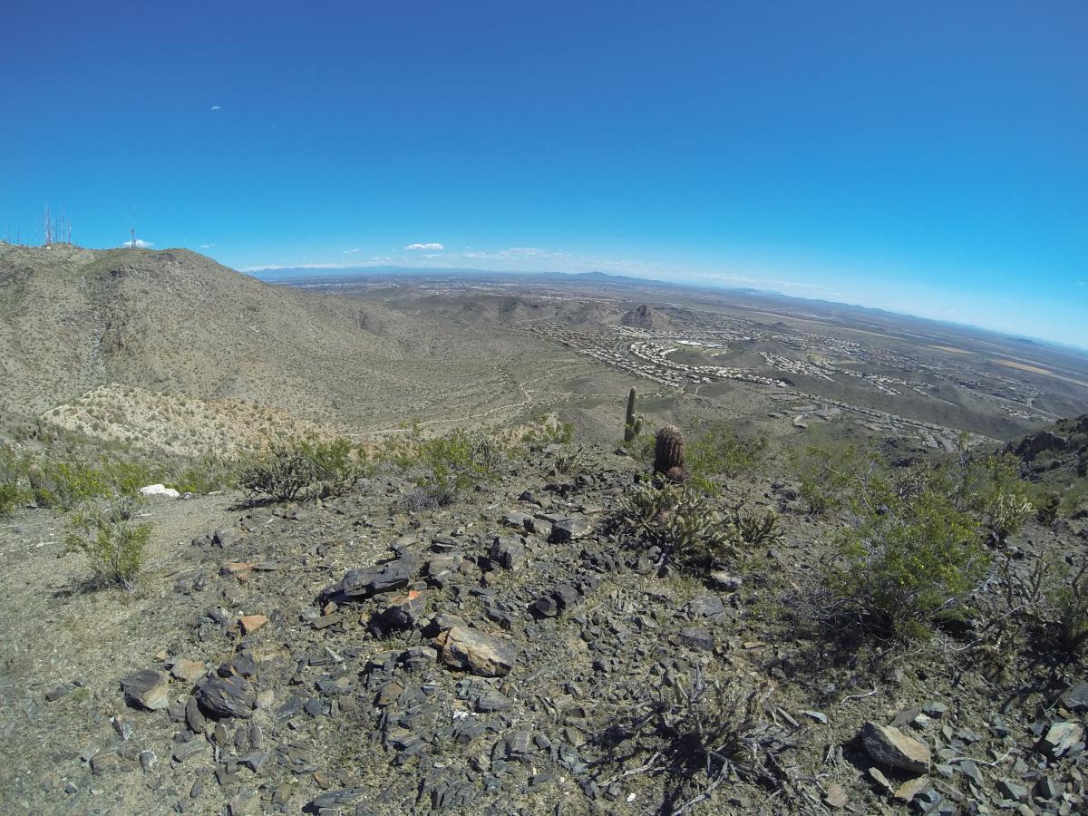 A panoramic view of a desert landscape, showcasing rolling hills, sparse vegetation, and a clear blue sky. In the foreground, rocky terrain is visible, with scattered shrubs and cacti. In the distance, a residential area is outlined against the horizon, with mountains faintly visible under a bright sky. South Mountain Park / National Trail mountain bike trail.