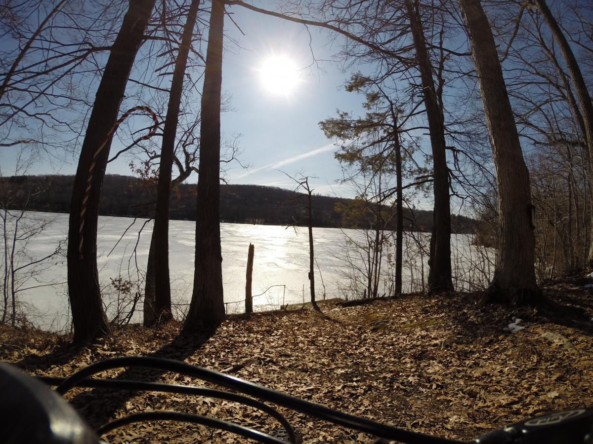 A scenic view of a frozen lake surrounded by trees under a bright sun, with fallen leaves on the ground in the foreground. The image captures a peaceful winter landscape, showcasing the natural beauty of the area. Kittatiny Valley State Park mountain bike trail.