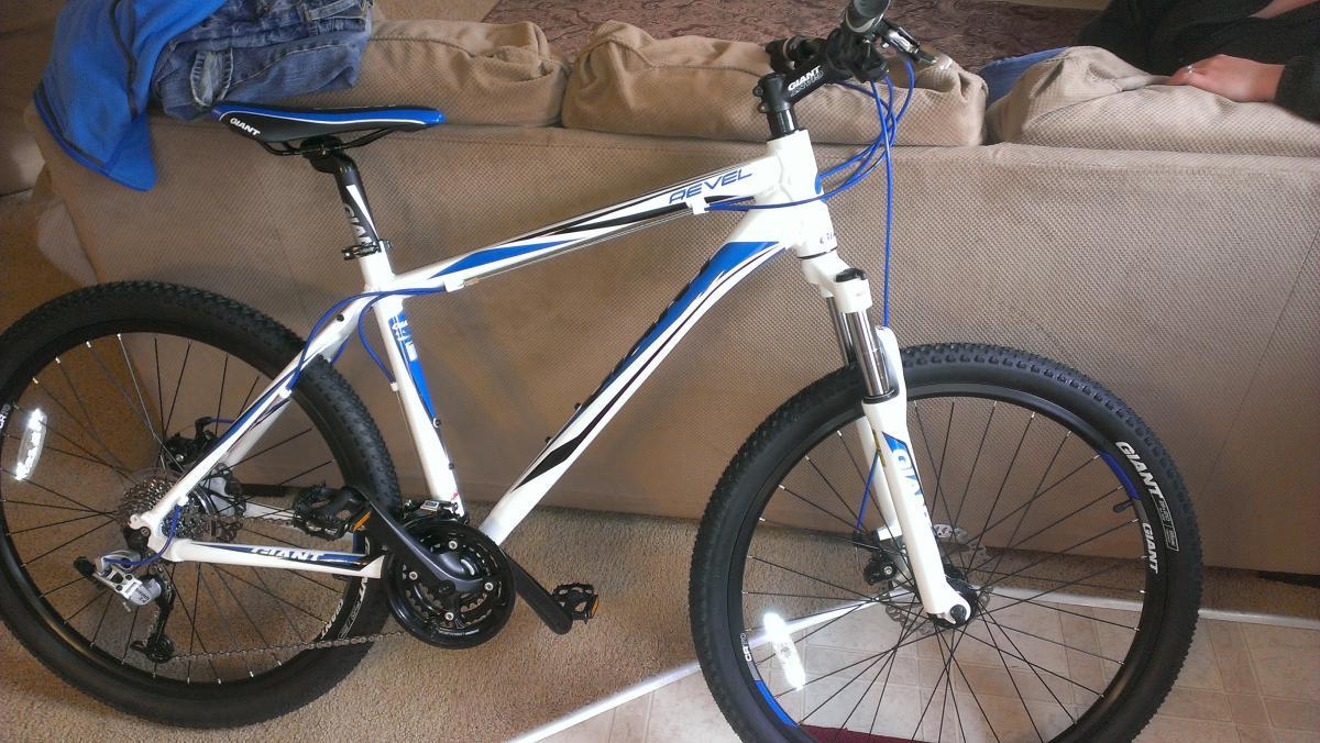Giant Revel 1: A white and blue mountain bike is leaning against a couch, showcasing its sleek frame and thick tires. The bike features a gear system and front suspension, suitable for off-road cycling. In the background, there are a pair of jeans and a blue shirt draped over the couch.