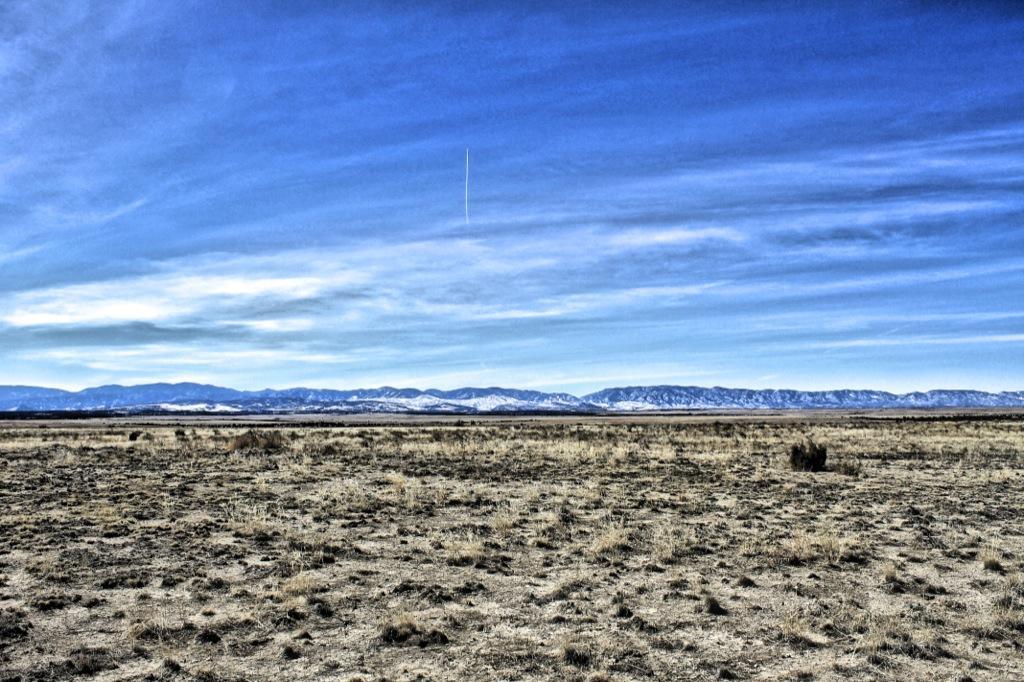 A panoramic view of a vast, open landscape featuring a dry, rocky terrain in the foreground, transitioning to distant mountain ranges capped with snow under a bright blue sky with wispy clouds. South Shore Lake Pueblo mountain bike trail.