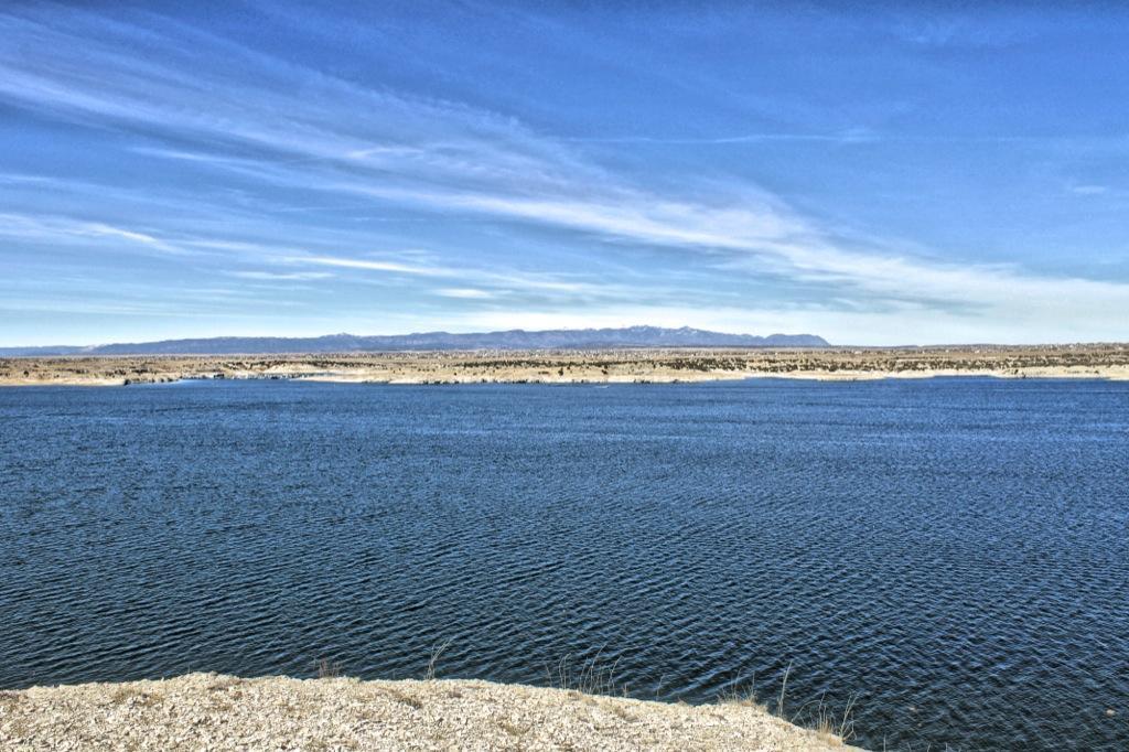 A serene landscape featuring a calm blue lake with gentle ripples, surrounded by arid land and distant mountains under a clear blue sky with wispy clouds. South Shore Lake Pueblo mountain bike trail.
