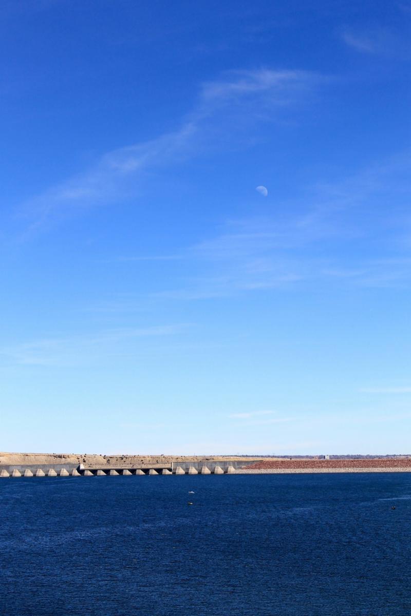 A serene landscape featuring a calm body of water under a clear blue sky, with a crescent moon visible in the upper left corner. In the background, a long structure extends across the water, resembling a dam or barrier, against a backdrop of lightly colored hills. The scene conveys a peaceful, natural setting. South Shore Lake Pueblo mountain bike trail.
