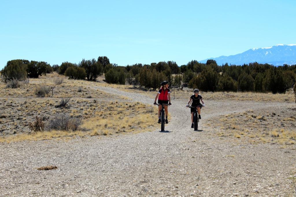Two cyclists riding on a gravel path through a semi-arid landscape, with scattered shrubs and distant mountains under a clear blue sky. South Shore Lake Pueblo mountain bike trail.