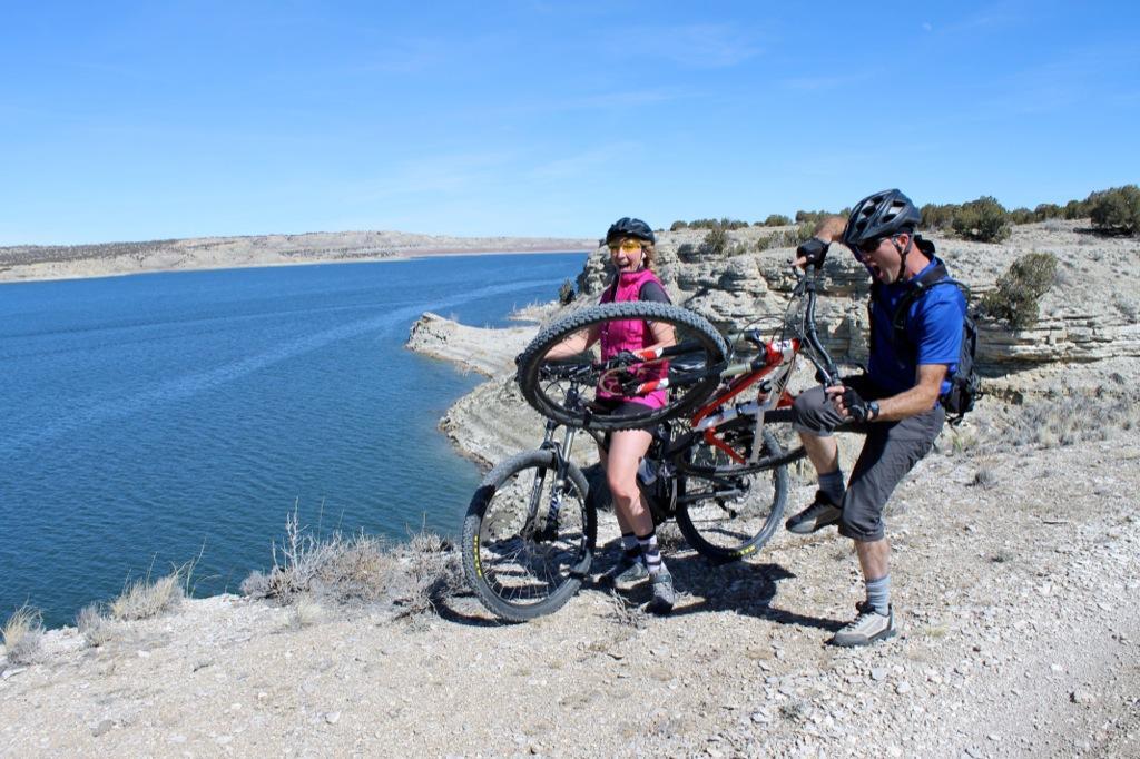 A man and a woman are on a mountain biking adventure near a lake. The woman, wearing a pink top and helmet, smiles as she holds her bike, while the man crouches nearby, examining the bike's wheel. The backdrop features a blue lake and rocky terrain under a clear sky. South Shore Lake Pueblo mountain bike trail.