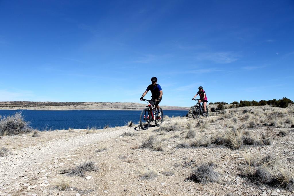 Two mountain bikers ride along a dirt trail by a blue lake, surrounded by sparse vegetation and rocky terrain under a clear blue sky. South Shore Lake Pueblo mountain bike trail.