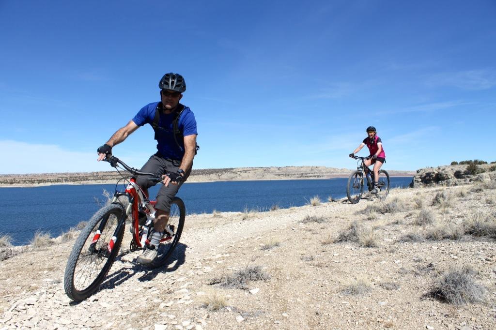 Two people biking along a dirt trail near a lake on a sunny day. The male cyclist, in a blue shirt and black shorts, is in the foreground, while a female cyclist, wearing a pink top and black shorts, follows behind. The landscape features dry terrain and clear blue skies. South Shore Lake Pueblo mountain bike trail.