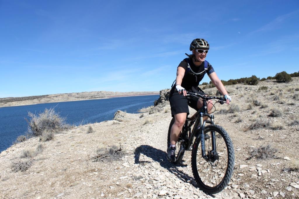 A woman riding a mountain bike along a rocky path near a lake, smiling. She is wearing a helmet and cycling gear, with sparse vegetation and a clear blue sky in the background. South Shore Lake Pueblo mountain bike trail.