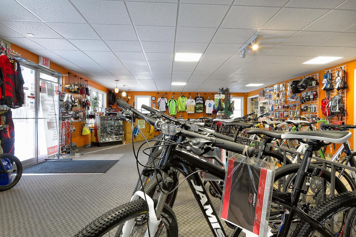 Interior of a bike shop showcasing a variety of bicycles lined up in the foreground. The walls are adorned with cycling apparel, accessories, and equipment, while the entrance features a glass door welcoming customers. Bright orange walls add a vibrant atmosphere to the space.