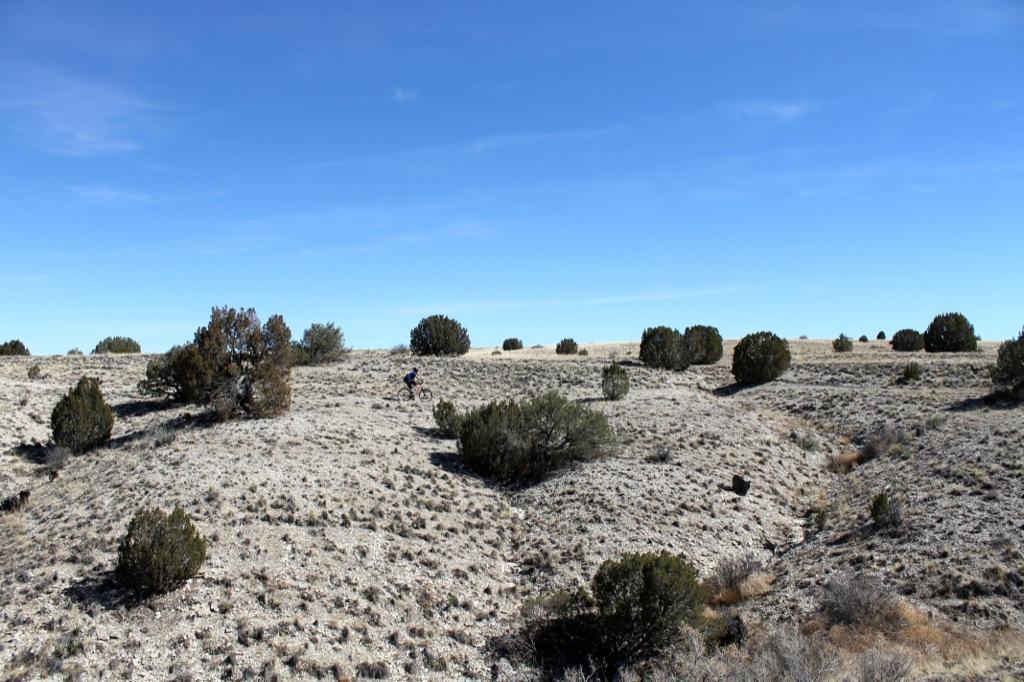 A mountain biker rides through a rocky, dry landscape dotted with small shrubs under a clear blue sky. The terrain features gentle hills and sparse vegetation typical of arid environments. South Shore Lake Pueblo mountain bike trail.