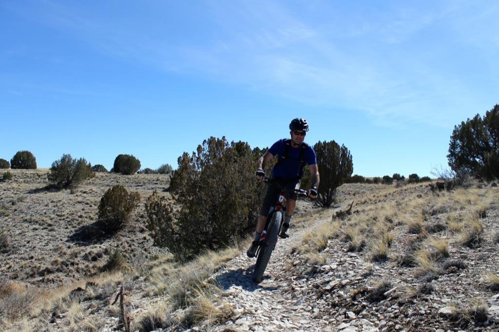 A person riding a mountain bike on a rocky trail in a rugged outdoor landscape. The scene features sparse vegetation, including small bushes and grass, under a clear blue sky. The cyclist is wearing a helmet and athletic clothing, with sunlight illuminating the path ahead. South Shore Lake Pueblo mountain bike trail.