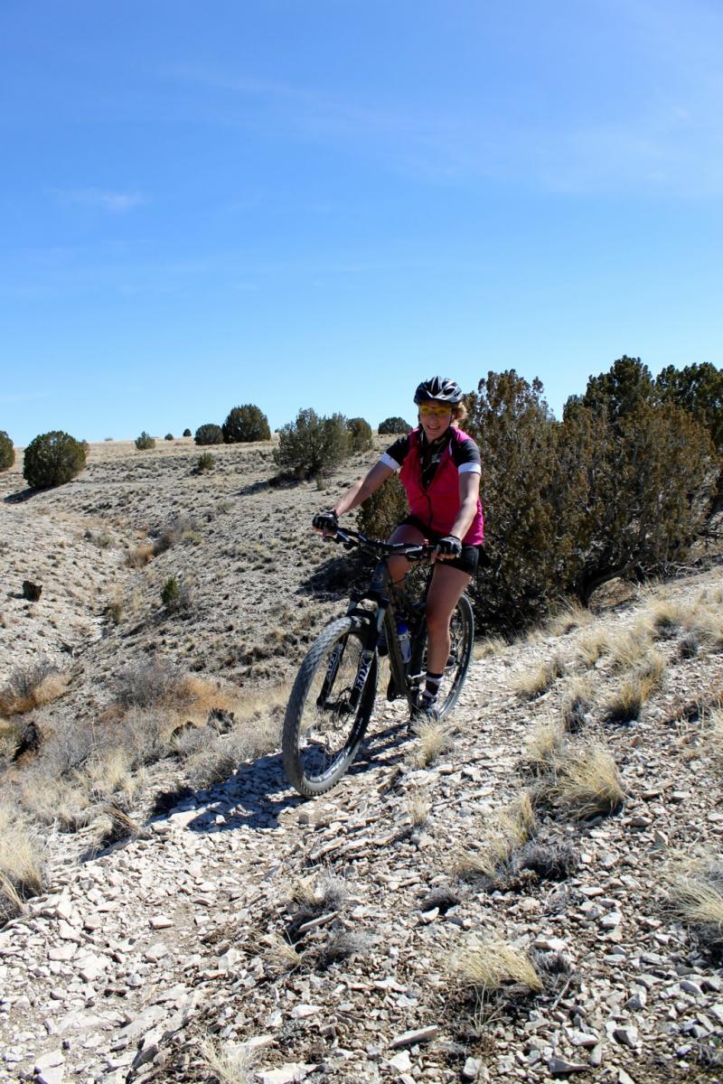 A person riding a mountain bike on a rocky trail in a dry, open landscape with sparse vegetation and blue skies in the background. South Shore Lake Pueblo mountain bike trail.