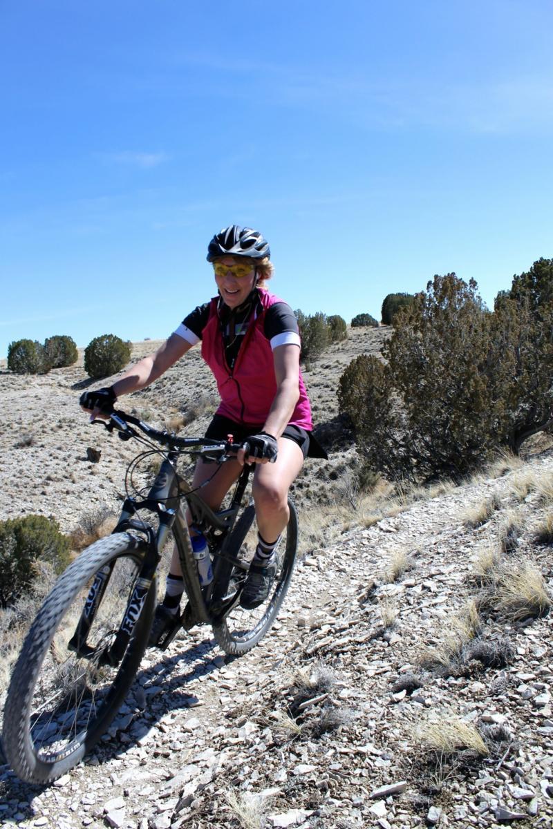 A smiling woman wearing a pink cycling vest and sunglasses rides a mountain bike on a rocky trail surrounded by sparse vegetation and blue skies. South Shore Lake Pueblo mountain bike trail.