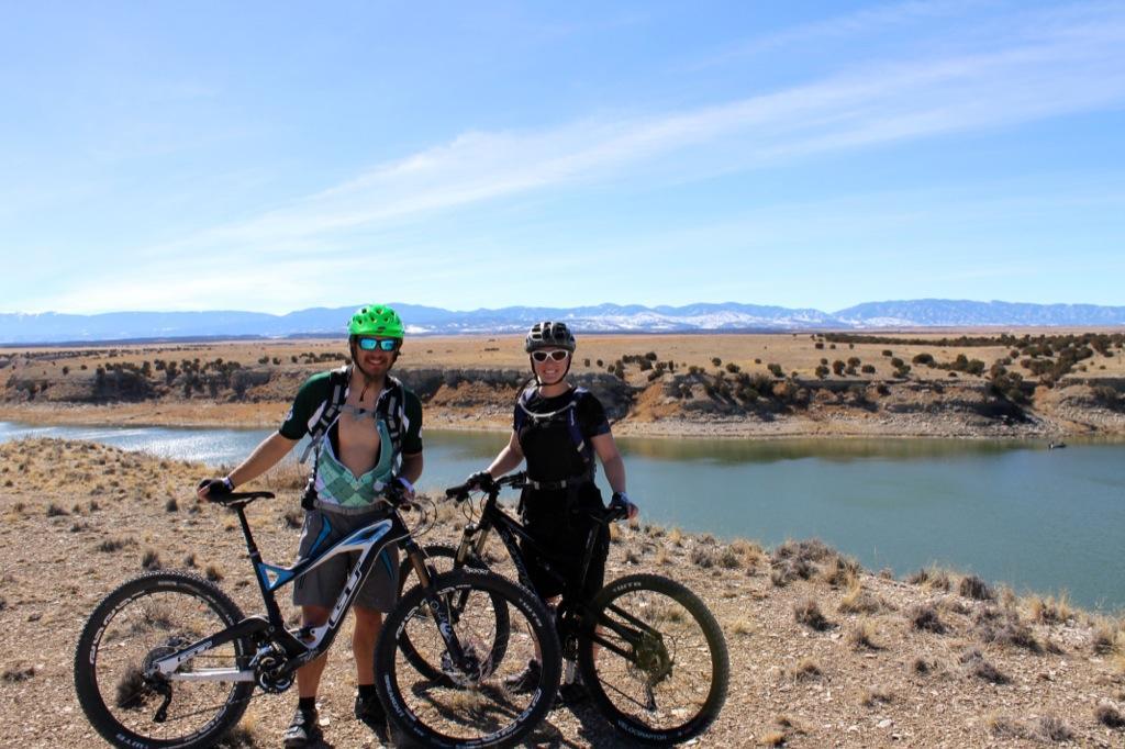 Two cyclists standing beside their mountain bikes near a body of water, surrounded by scenic hills and mountains. The sky is clear and blue, indicating a sunny day. One cyclist is wearing a green helmet and a short-sleeved jersey, while the other is in a black outfit with a helmet. They appear to be enjoying a break during their biking adventure. South Shore Lake Pueblo mountain bike trail.