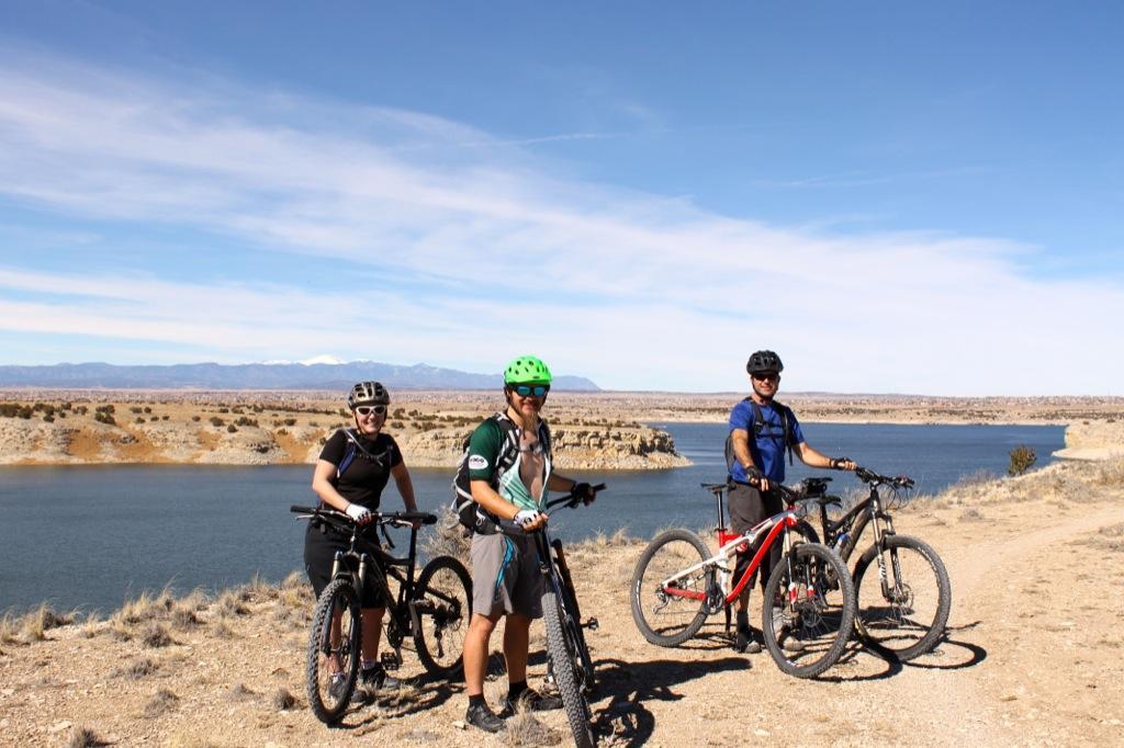 Three mountain bikers pose with their bikes near a lake, surrounded by a scenic landscape featuring rolling hills and a clear blue sky. The group includes two men and one woman, all wearing helmets and cycling gear, with distant mountains visible in the background. South Shore Lake Pueblo mountain bike trail.
