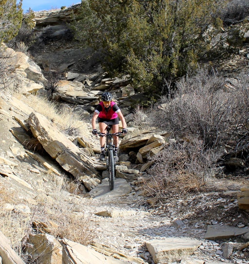 A mountain biker navigating a rocky trail surrounded by sparse vegetation and rocky terrain, wearing a helmet and a pink cycling jersey. The cyclist is focused on balancing as they ride over the uneven ground under a clear sky. South Shore Lake Pueblo mountain bike trail.