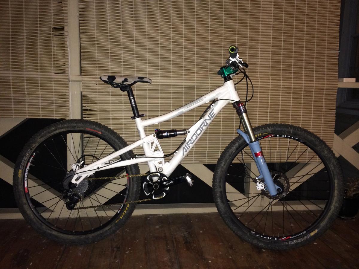 Airborne Zeppelin Elite: A white mountain bike with a blue suspension fork, showcasing knobby tires and a sleek frame. The bike is positioned on a wooden floor with a textured backdrop featuring horizontal blinds.