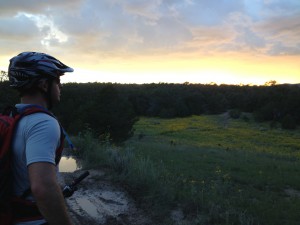 A cyclist stands on a dirt path, gazing at a vibrant sunset over a green landscape dotted with yellow wildflowers. The sky is filled with soft, colorful clouds, and the cyclist is wearing a helmet and sport attire, with a mountain bike resting by their side.