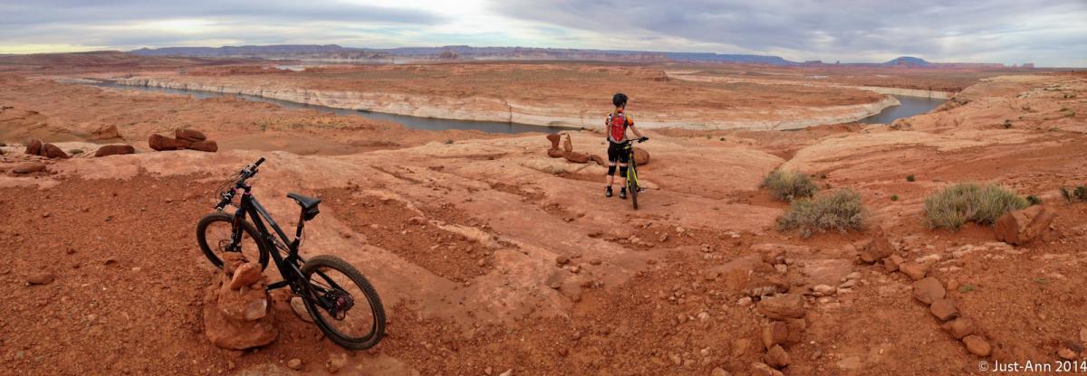A mountain biker standing on a rocky ledge overlooking a vast desert landscape with a winding river below. A black mountain bike is parked nearby, surrounded by red rock formations and sparse vegetation under a cloudy sky. Page Rimview mountain bike trail.