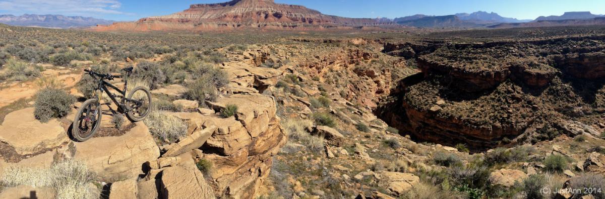 A mountain bike is parked on a rocky ledge overlooking a vast canyon landscape with shrubs and desert vegetation. In the background, distinct, layered rock formations and distant mountains are visible under a clear blue sky. J.E.M. Trail mountain bike trail.