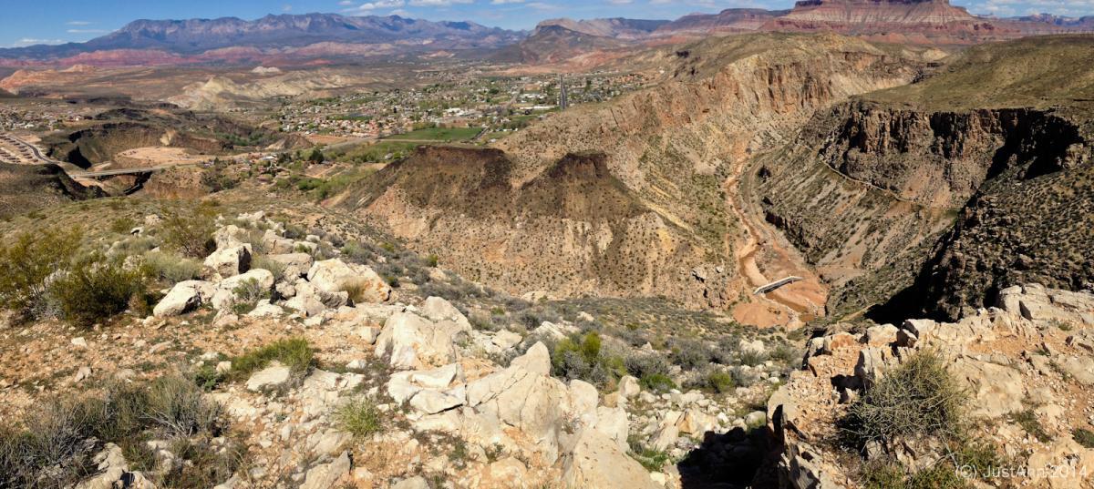 A panoramic view of a rugged landscape featuring rolling hills, rocky cliffs, and a winding river. The foreground includes rocky terrain and sparse vegetation, while the background showcases distant mountains under a clear blue sky. A small town is visible nestled in the valley below, surrounded by green fields and roads. J.E.M. Trail mountain bike trail.