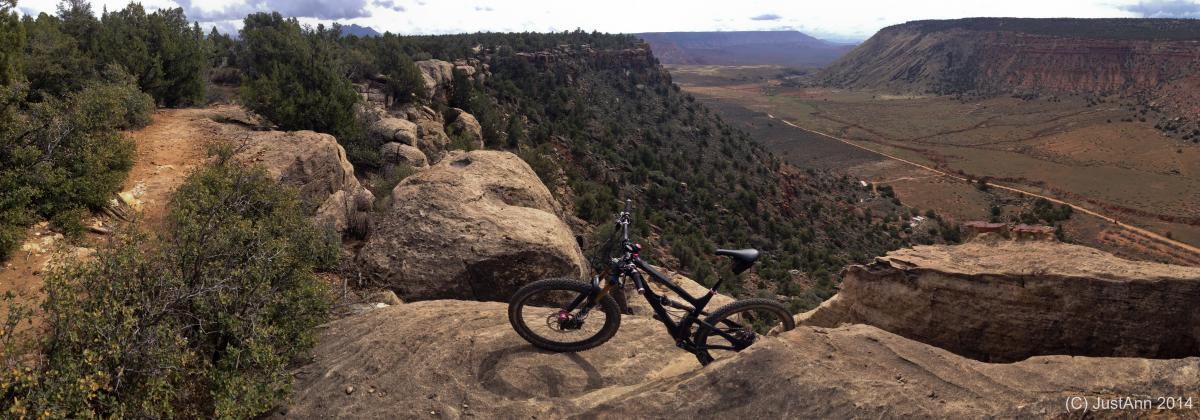 A mountain bike resting on a rocky outcrop overlooking a vast, green valley. Pine trees surround the area, and the landscape features dramatic cliffs in the distance under a partly cloudy sky. Guacamole Mesa mountain bike trail.