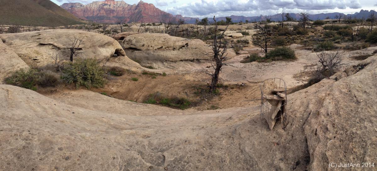A panoramic view of a rugged desert landscape featuring sandy rocky terrain and sparse vegetation. In the foreground, a bent wire structure is partially covered with fabric, while in the background, distant mountains and cloudy skies are visible, creating a dramatic backdrop. Guacamole Mesa mountain bike trail.