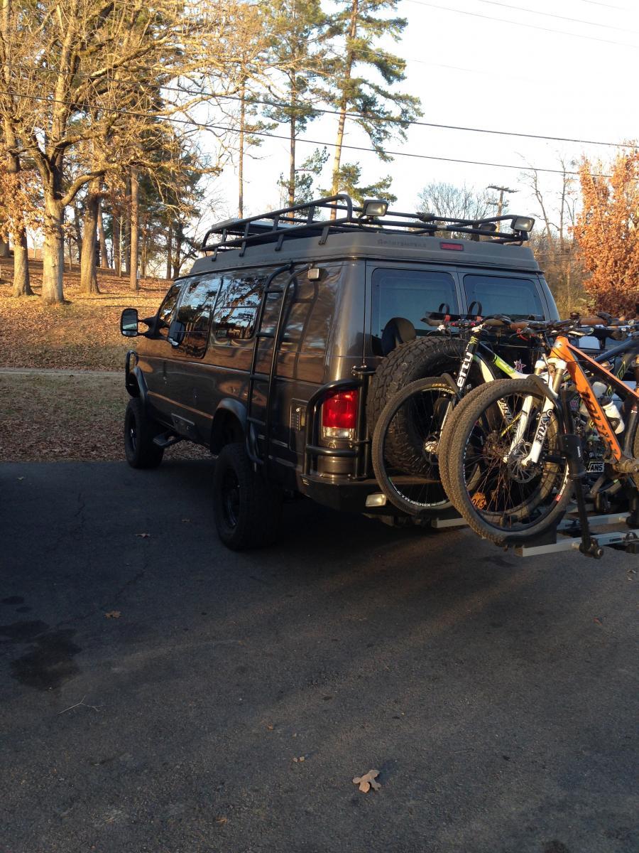 A dark-colored van parked on a paved area, equipped with a bicycle rack holding three bicycles. The background features trees with autumn leaves and power lines, indicating a natural setting. Spadra Creek Nature Trail mountain bike trail.