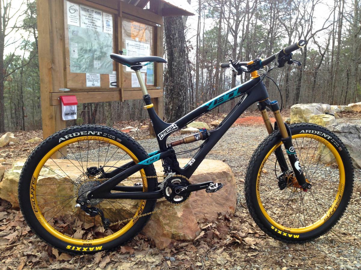 A black mountain bike with yellow Maxxis Ardent tires stands on a rocky surface in a forested area. Behind the bike, there is a wooden information board displaying maps and notices. The background features tall trees and a gravel path covered with fallen leaves. Coldwater Mountain mountain bike trail.