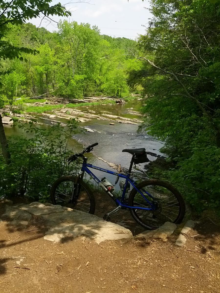 Kona Cinder Cone: A blue mountain bike leaning on a rocky path beside a serene river, surrounded by lush green trees and foliage under a clear sky.
