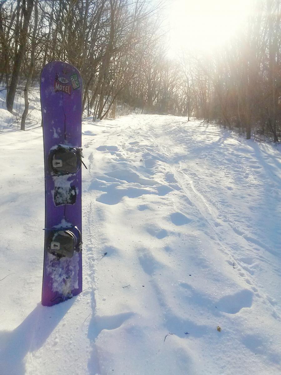 A purple snowboard leaning against a snow-covered trail, surrounded by bare trees. The sun shines brightly in the background, illuminating the snowy landscape. Raceway Woods mountain bike trail.