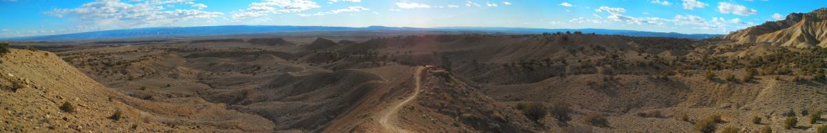 A panoramic view of a desert landscape featuring rolling hills and ridges, with a winding dirt path cutting through the terrain. The sky is partly cloudy, and the distant horizon showcases a blend of blue and light clouds. The scene conveys a sense of vastness and natural beauty in an arid environment. Zippety Do Dah mountain bike trail.
