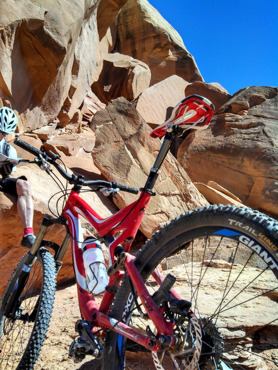 Specialized Stumpjumper FSR Pro: A close-up view of a red mountain bike parked on rocky terrain with a clear blue sky overhead. In the background, a person wearing a helmet and cycling attire sits on a rock, appearing to look at a map or GPS device. The bike features a water bottle holder and thick, textured tires designed for trail riding.