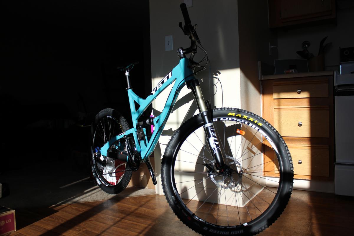 Yeti SB-75: A turquoise mountain bike with thick tires, positioned in a sunlit area, showcasing its frame and details against a backdrop of wood flooring and kitchen cabinetry.