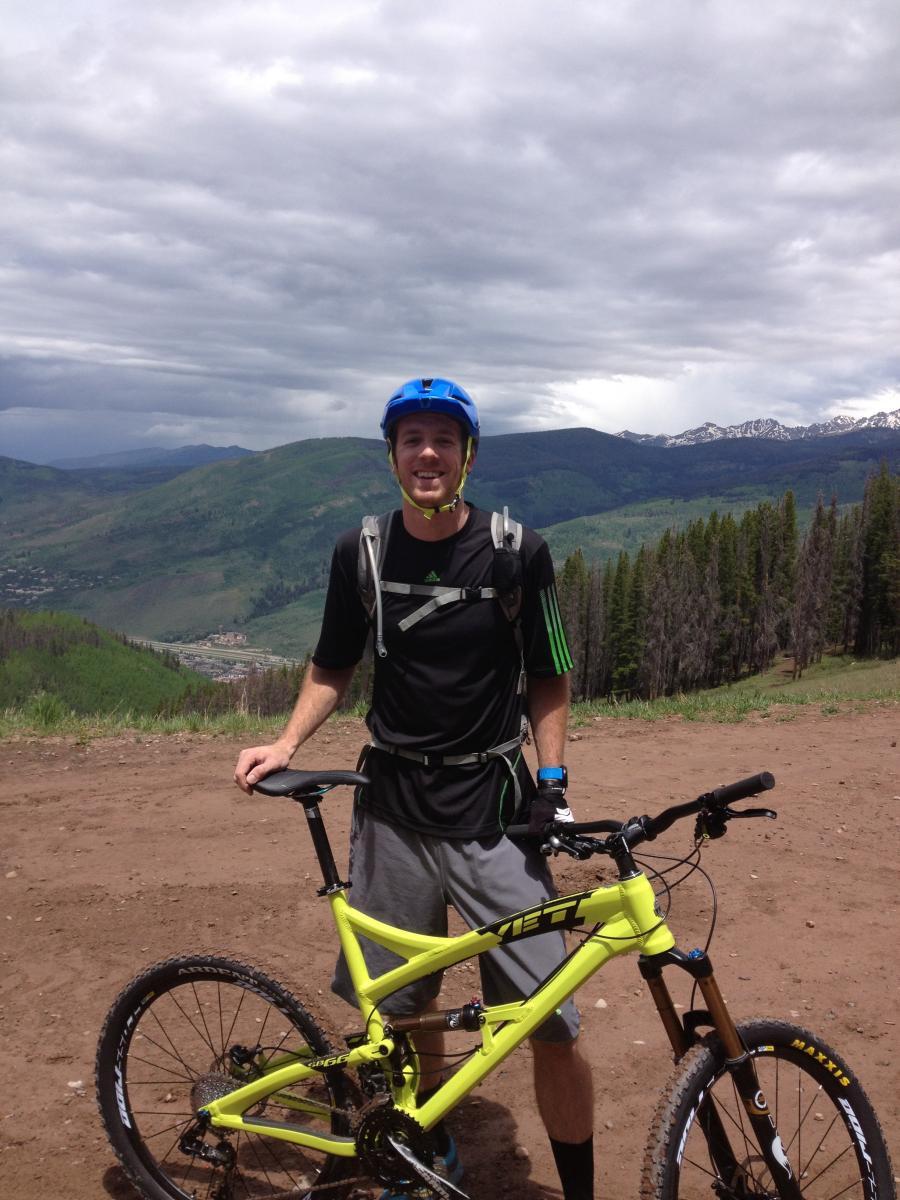Yeti SB66: A cyclist wearing a blue helmet and a black athletic shirt stands next to a bright yellow mountain bike on a dirt trail. In the background, a scenic view of green hills and snow-capped mountains under a partly cloudy sky is visible.
