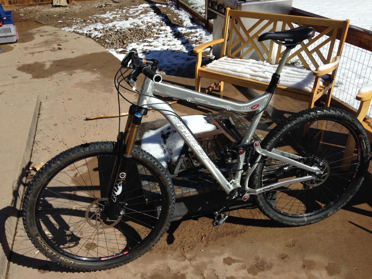 Niner RIP 9: A silver mountain bike with thick tires, positioned on a sandy surface. The bike features a suspension system and a sleek design, with the handlebars and wheels clearly visible. In the background, there’s a wooden bench partially covered in snow, and some scattered stones.
