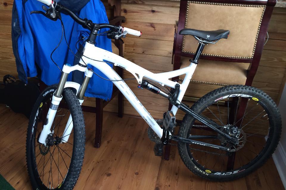 Titus Moto Lite: A white mountain bike with black accents, featuring front suspension, standing on a wooden floor. In the background, there is a blue jacket draped over a chair and a black bag partially visible.