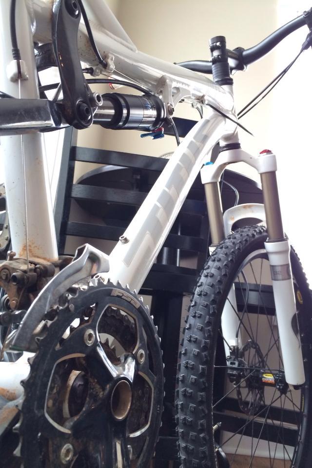 Titus Moto Lite: Alt text: Close-up view of a white mountain bike, focusing on the chainring, rear suspension, and front fork. The background includes a black chair, indicating an indoor setting. The bike shows signs of use with some dirt on the components.