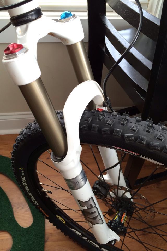 Titus Moto Lite: Close-up image of a mountain bike's front fork, featuring a white Fox Racing Shox suspension with red and blue adjustable knobs on top. The bike's knobby tire is partially visible, along with a wooden floor and a chair in the background.
