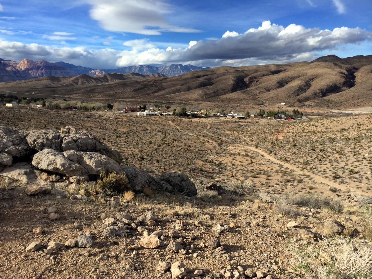 A scenic view of a desert landscape featuring rocky terrain and sparse vegetation, with distant hills and mountains under a partly cloudy sky. A small town is visible in the valley below, surrounded by the expansive desert. Blue Diamond mountain bike trail.