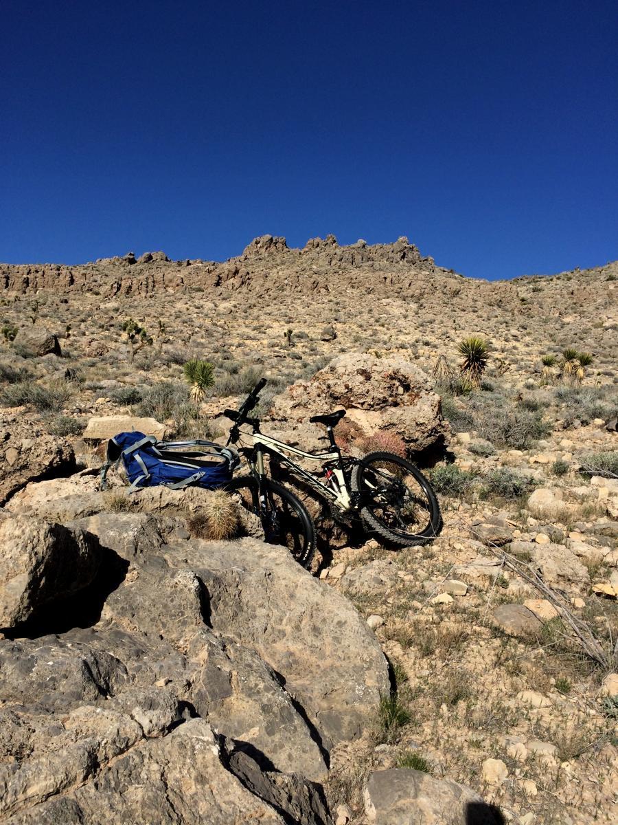 A mountain bike and a blue backpack resting on rocky terrain under a clear blue sky, with rugged hills in the background. Blue Diamond mountain bike trail.