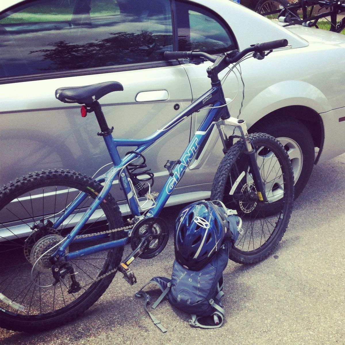 Giant Yukon: A blue mountain bike leaning against a silver car, with a black helmet and a gray backpack on the ground beside it. The bicycle features knobby tires and a prominent brand logo.