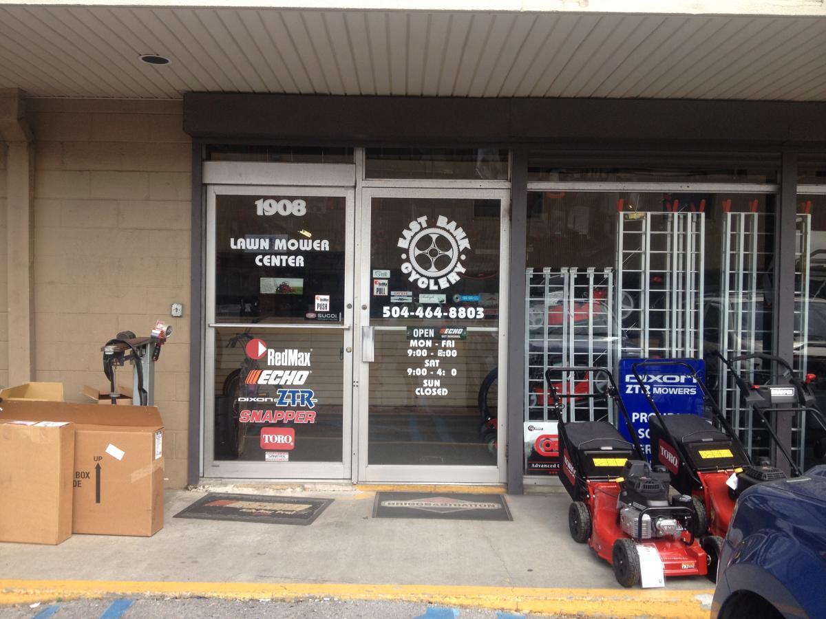 Alt text: Exterior view of a lawn mower center featuring a glass door with a sign displaying "LAWN MOWER CENTER" and various brand logos. On the sidewalk, there are boxes and lawn mowers on display, along with contact information for the business.