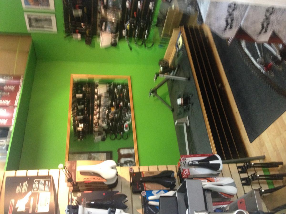 A brightly colored bicycle shop interior featuring a green wall with various cycling accessories displayed. The image includes shelves of bike components, including saddles and handlebars, as well as a wooden counter with additional items. The setting conveys a lively atmosphere for cycling enthusiasts.