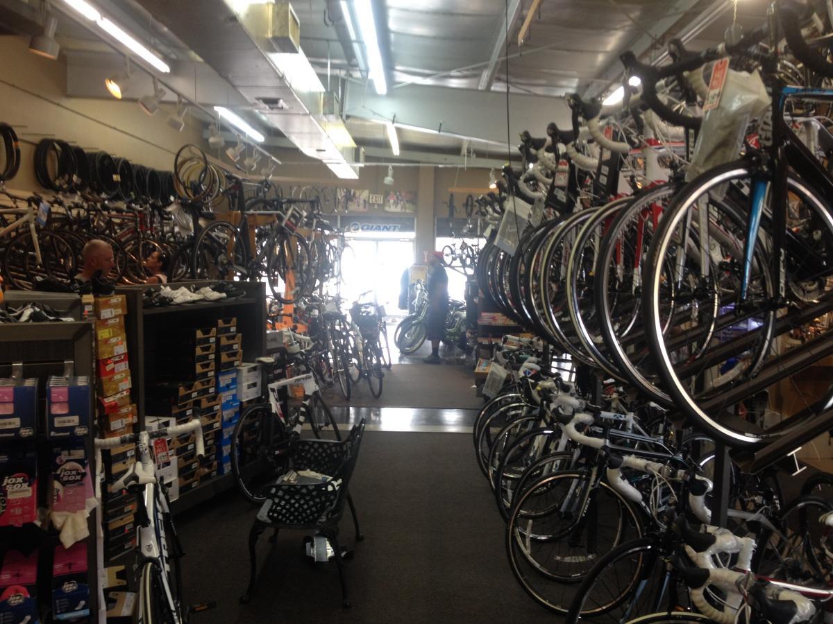 A brightly lit bike shop filled with various bicycles and cycling accessories. The interior features numerous bikes displayed on racks and hanging from the walls. Shelves are stocked with bike gear and shoes, while a couple of customers are browsing near the entrance.