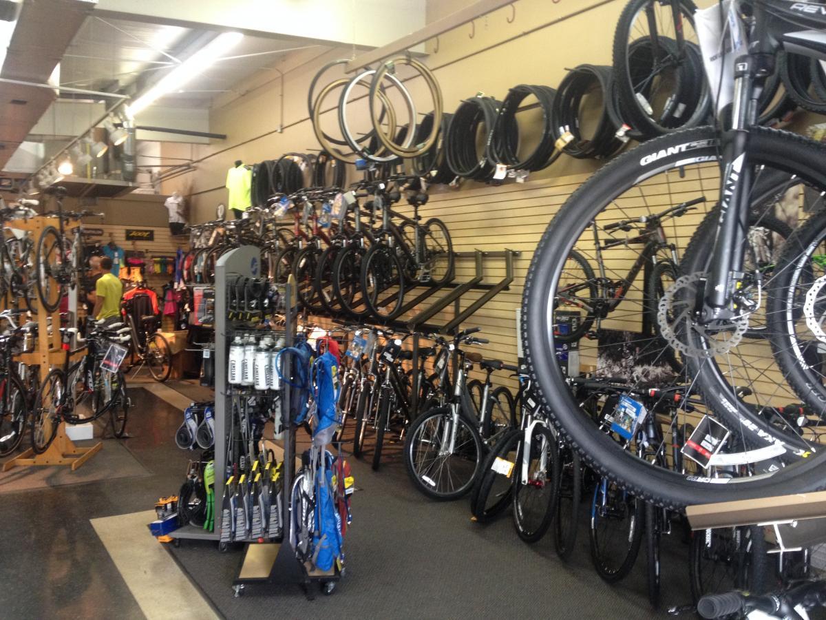 A spacious bicycle store filled with various models of bikes, accessories, and gear. The interior features rows of bicycles hanging on the walls, along with racks displaying tires and cycling apparel. A display stand showcases water bottles and other biking accessories in the foreground, while customers browse the selection in the background.