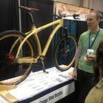 A person in a green shirt and another individual with long hair are engaged in conversation while examining a unique wooden bicycle on display at a trade show booth. The bicycle features a sleek design with wooden frame components, and it stands on a table alongside brochures and information about the brand "Connor Wood Bicycles." The booth is decorated with a banner that reads "Sojourner Cyclery."