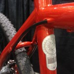 Close-up view of a shiny red bicycle frame featuring a metallic badge and cable routing. The frame includes thick, knobby tires in the background, indicating a design suited for off-road riding.